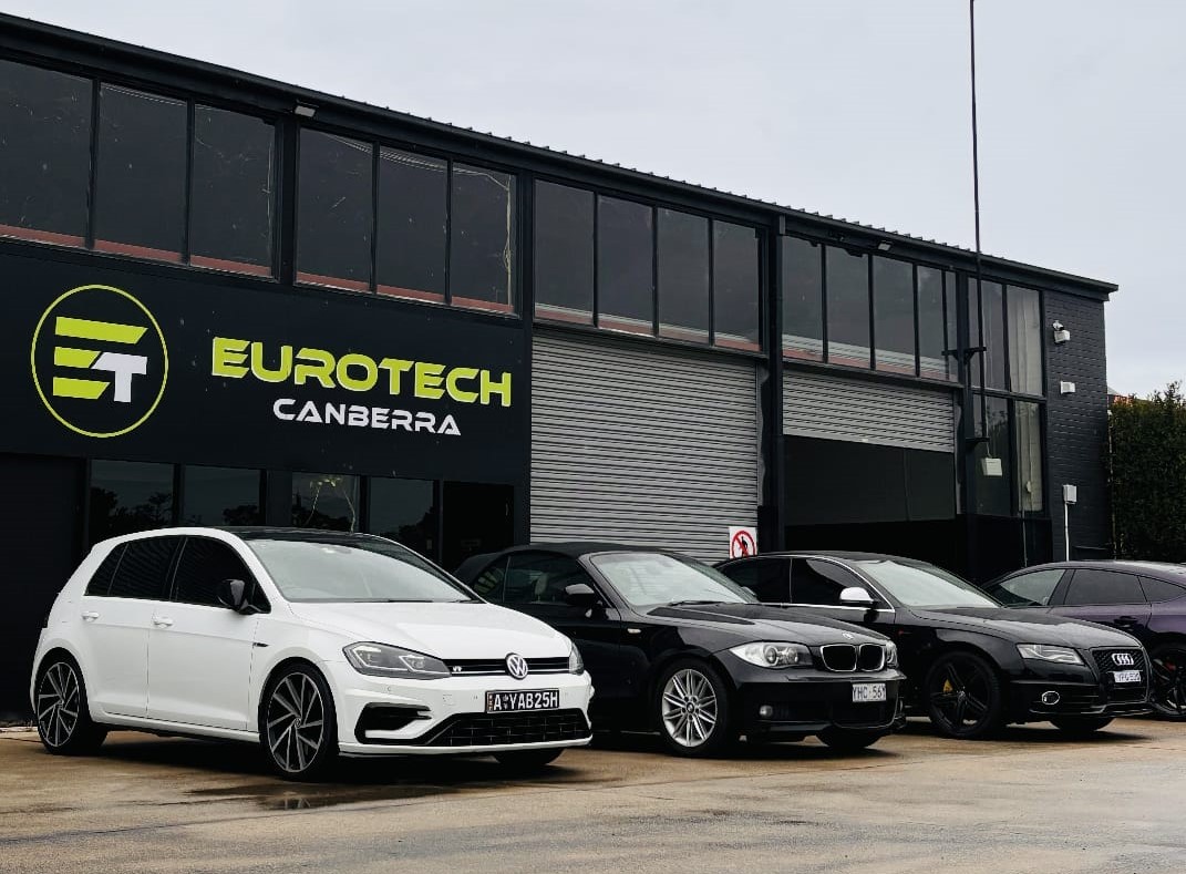 A modern automotive workshop named "Eurotech Canberra" with a row of European cars parked in front, including a white Volkswagen, a black BMW, and two black Audis. The building has large glass windows and a prominent sign with the Eurotech logo.